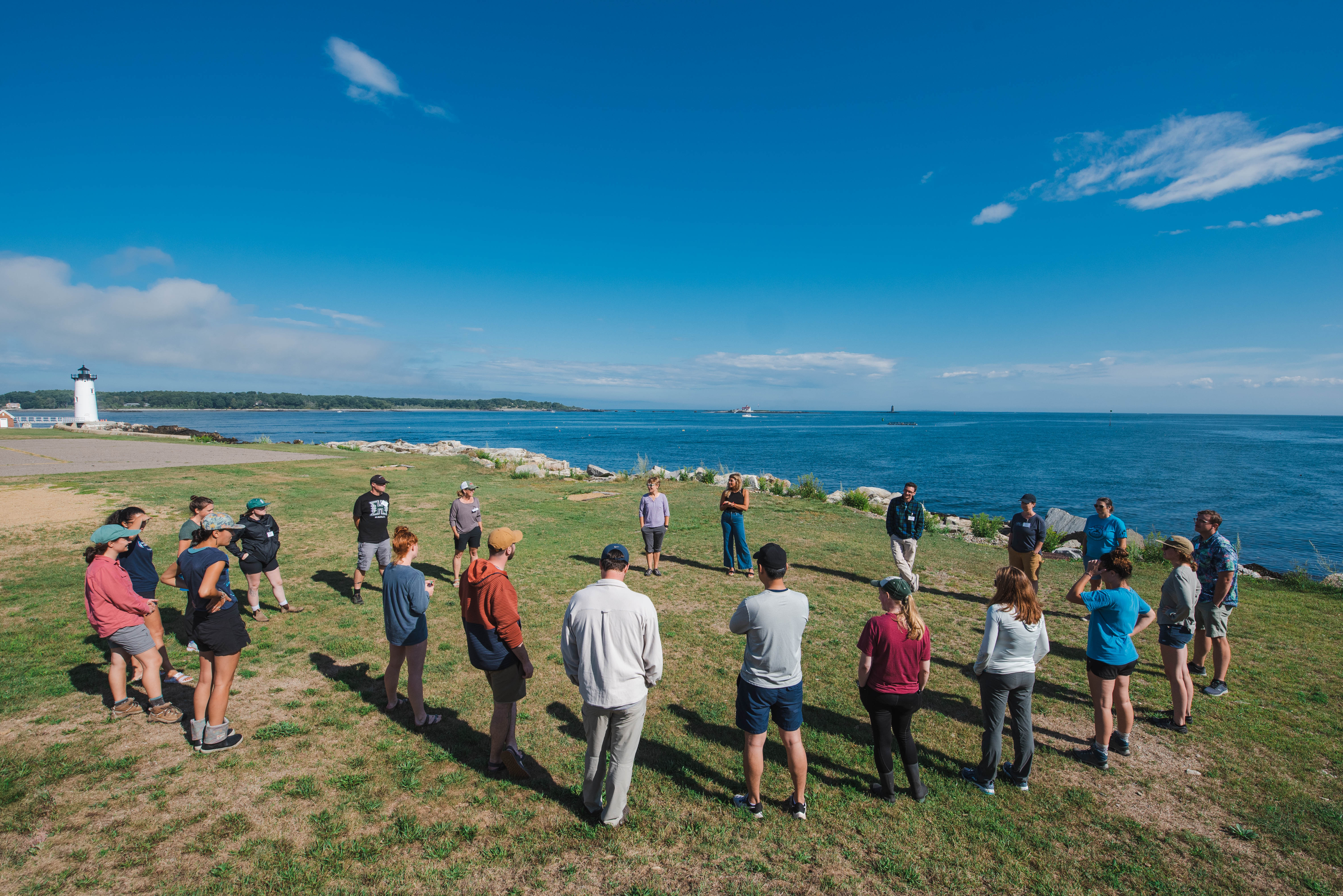 A group of CoastWise participants stands in a circle on New Hampshire's rocky shoreline, overlooking the ocean.