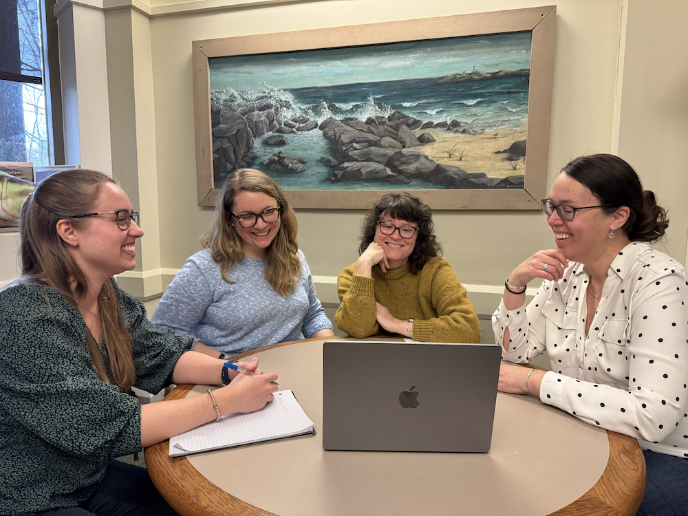 Four women sit at a desk together, looking at a laptop and smiling.