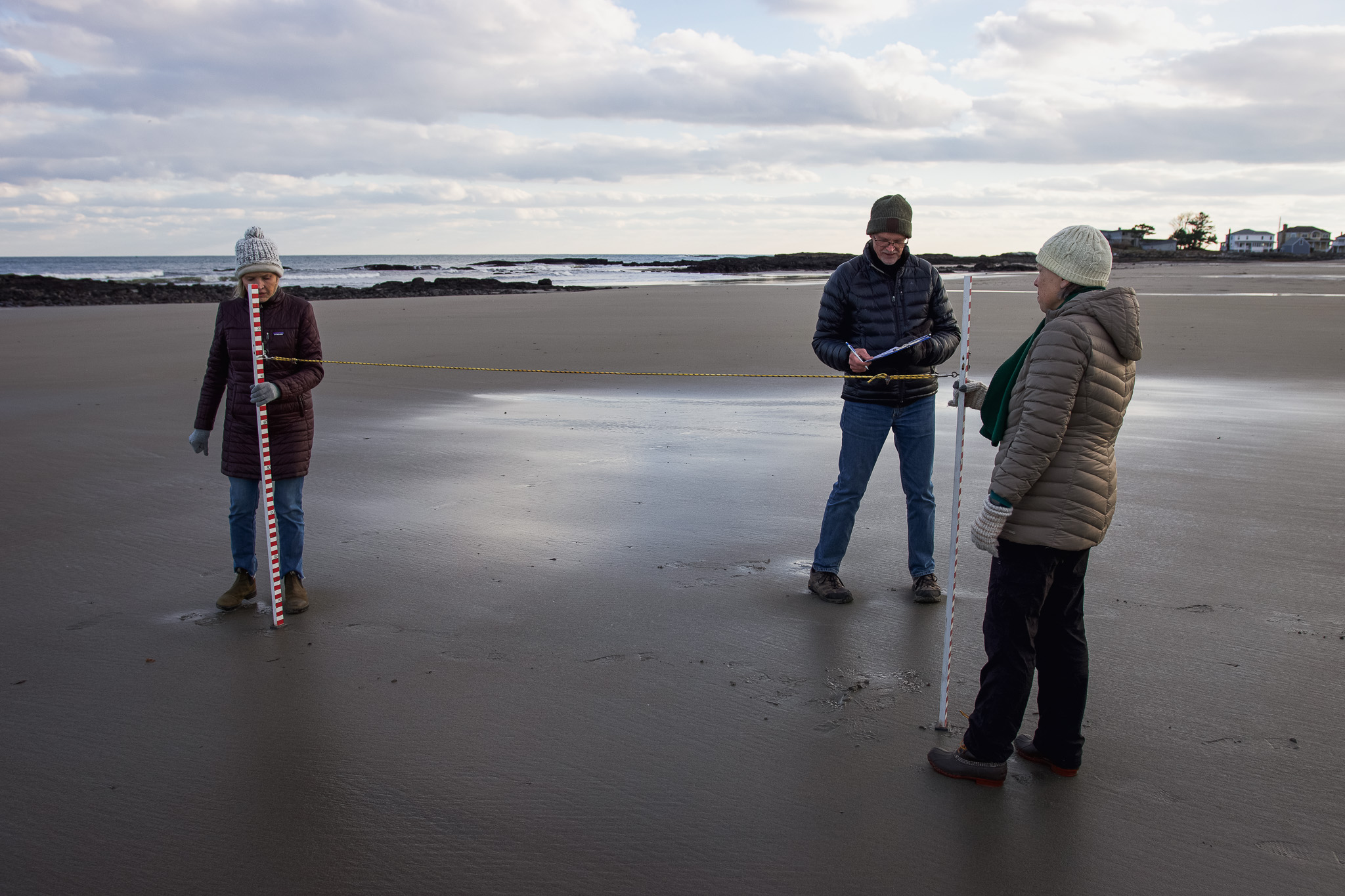 A group of 3 volunteers stand with two measuring poles and a clipboard on the beach. 