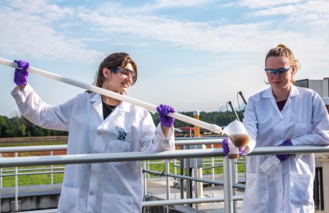 two women in white lab coats use a long pole with a cup on the end to sample wastewater at a treatment facility in New Hampshire