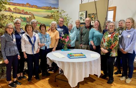 Fifteen smiling adults stand in a semi-circle behind a table with a white tablecloth on which sits a celebratory cake commemorating their graduation from UNH Marine Docent Program