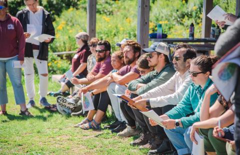 CoastWise participants outside on a sunny summer day sit in a line and observe a presentation during a professional development session