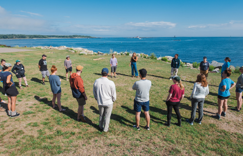 group of about twenty people from the New Hampshire CoastWise program stand in a circle on a grassy lawn overlooking the mouth of the Piscataqua River in New Castle, NH with the white Fort Constitution lighthouse in the background