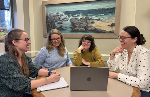 Four women sit at a desk together, looking at a laptop and smiling. 