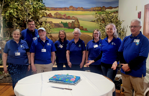 UNH Marine Docent Class of 2025 group photo of 8 adults wearing their blue docent polos standing behind a round table with their celebratory cake displayed