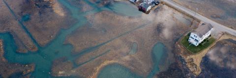 An aerial view of a marsh at high tide and a house with a thin ring of lawn separating it from the waterline against a road through the marsh