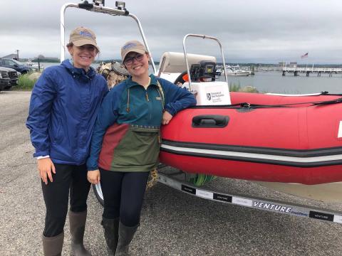 Kayla Tozier and researcher Cate pose in front of a red inflatable boat on a trailer near the shoreline