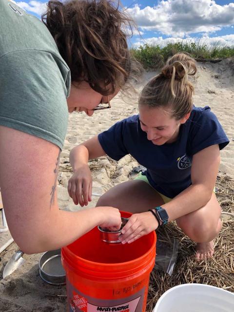 Kayla and Cate bend down over a bucket on a sandy beach while they sieve sand into the bucket to take sand samples for microplastics