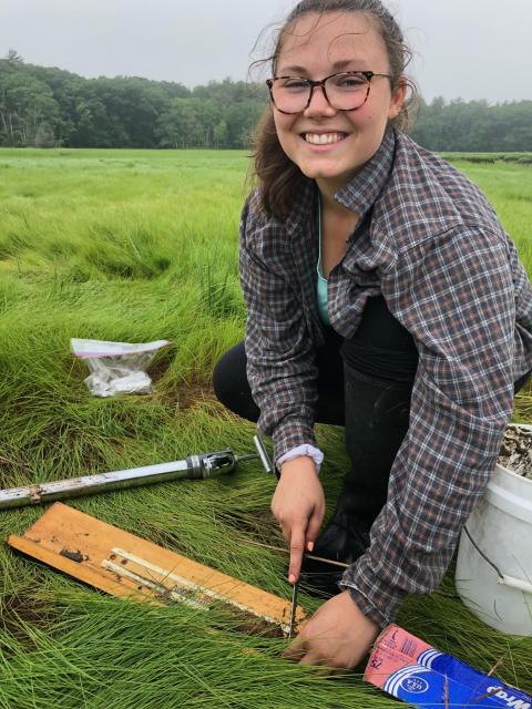 Kayla Tozier poses while cutting a sediment core to length while kneeling in a salt marsh