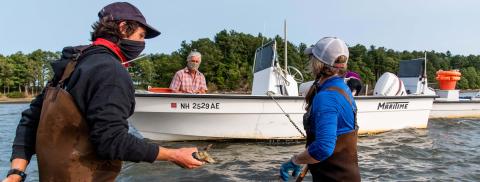 An oyster farmer, standing in waist deep water in waders, speaks to a researcher in a boat and another researcher also standing in the water in Great Bay, NH
