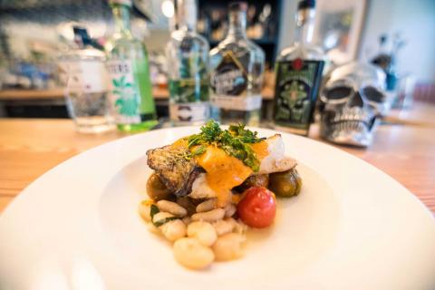 A dish of sautéed striped bass with beans and orange sauce on a white plate in front of bottles of gin and a silver decorative skull on a bar