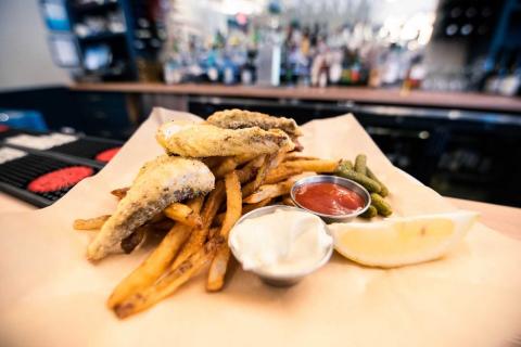 A photograph of fried striped bass on a bed of French fries with pickles, mayonnaise, ketchup, and a lemon in front of a stocked bar
