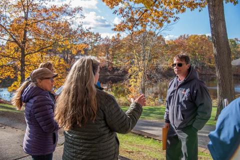 Three people stand talking in a park in front of a pond surrounded by trees with fall foliage