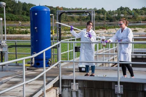 A researcher pours a beaker of dark liquid into another beaker held by a second researcher at a Wastewater Treatment Facility