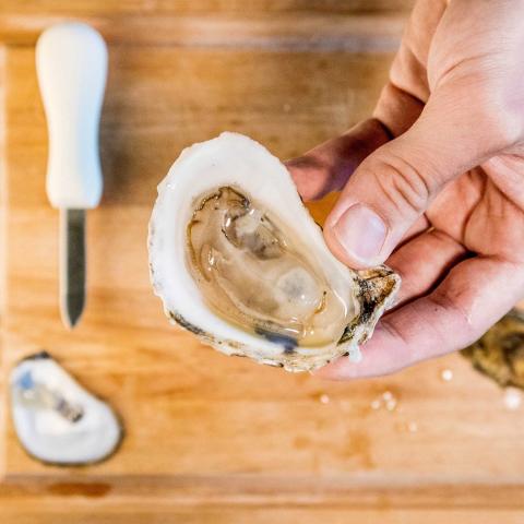 A shucked oyster is held over a wooden cutting board with an oyster knife and the other half of the shell below