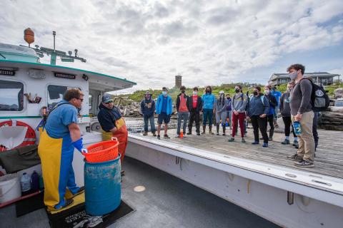 students stand on a dock at Appledore Island facing a lobster boat with two lobstermen wearing foul weather pants