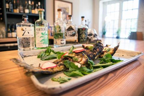 A photograph of a whole grilled striped bass with greens and radishes in a sheet pan on a bar in a restaurant in front of bottles of gin