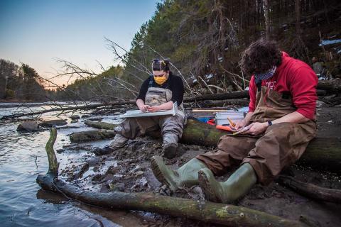 Two researchers sit on the side of a river measuring and counting smelt
