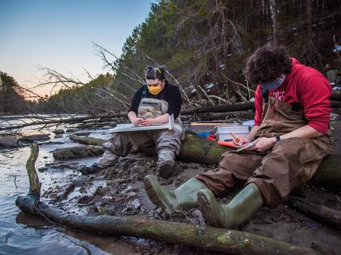 Two researchers sit on the side of a river measuring and counting smelt