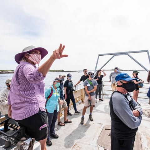 people stand on the back of a boat as a woman points while lecturing to the people