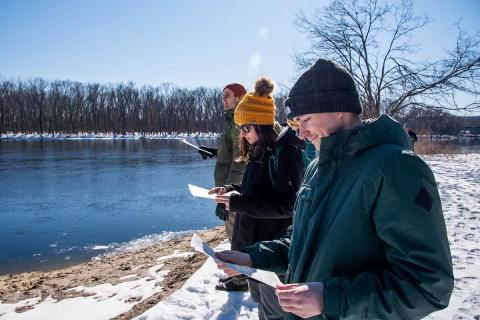 Three people in warm winter clothes stand on the bank of a river holding papers in their hands