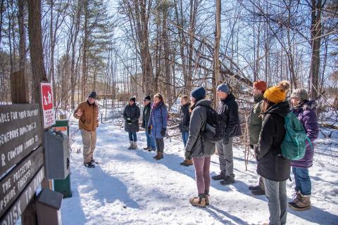 A group of people in warm winter clothes stand in a snowy forest in front of a sign while a man talks to them