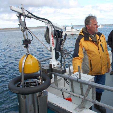 a man in a yellow rain jacket stands next to a barotrauma device hanging over the side of a boat over the water