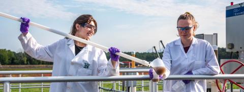 two women in white lab coats use a long pole with a cup on the end to sample wastewater at a treatment facility in New Hampshire