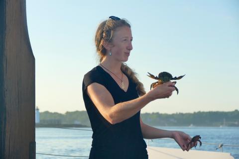 Emily Burke stands holding two green crabs in front of a view of water with trees behind under a blue sky