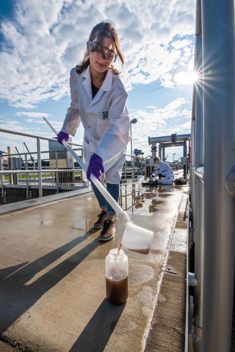 A researcher wearing a white lab coat, blue gloves, and safety goggles pours a beaker with brown liquid on the end of a stick into a bottle. Another researcher in the background preps samples.