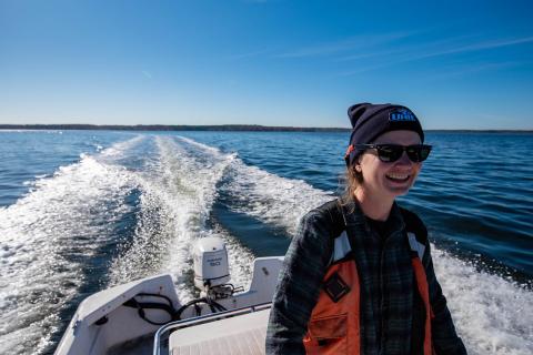 Researcher Kelsey Meyer wearing a life jacket, hat, and sunglasses, drives a boat in Great Bay with foam trailing behind in blue waters