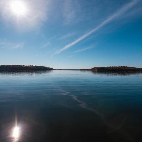 A photo of Great Bay, calm water reflects a blue sky with small streaks of clouds and a bright sun above a faraway tree-lined shoreline