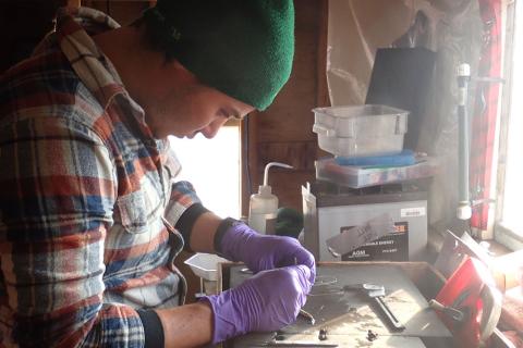 researcher Nate Hermann works with a sample while wearing purple nitrile gloves, a flannel shirt and a green beanie