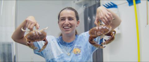Jillian Robillard, smiling, holds two Jonah Crabs (reddish crabs about 8 inches across) in her facility, wearing a blue tie dye shirt with the Southern Maine Crabs logo on it