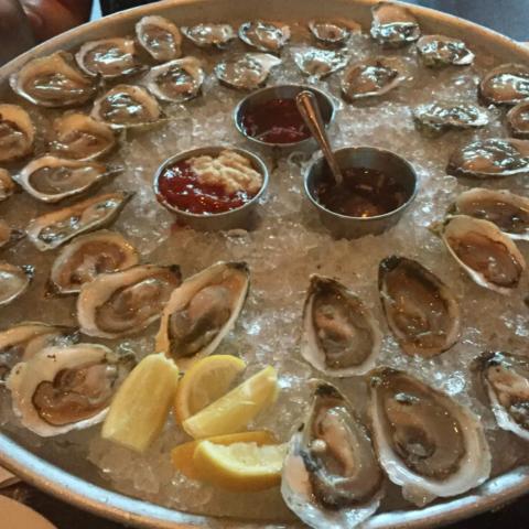 A photograph of dozens of oysters on the half shell on a bed of ice with lemon wedges and small dishes of sauces