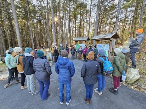 CoastWise participants stand outside in a circle at the start of the All Persons Trail in Manchester with tall trees surrounding