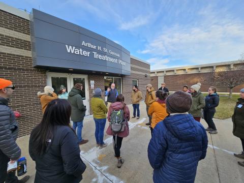 CoastWise participants outside on a chilly spring day stand in a circle in front of a water treatment plant entrance