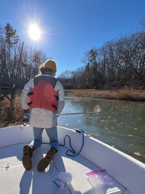 Gabby Deangelis kneels on the bow of a boat, with a skimmer to collect foam samples.