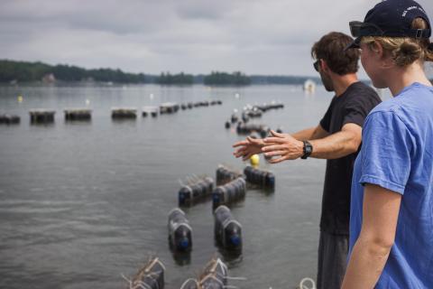 Emma stands on a vessel with an oyster farmer, looking out on his farm. 