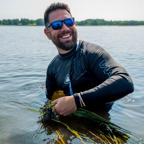 Trevor Mattera standing in the water holding a bundle of eelgrass and smiling at the camera. 