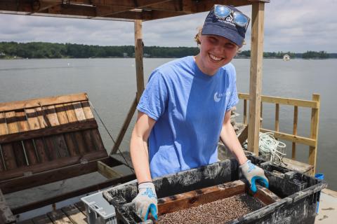 A student stands on an oyster farm, smiling and holding a bin with oyster spat. 
