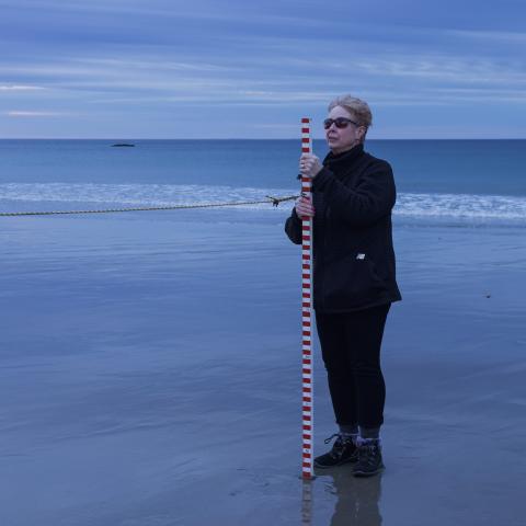 Deborah holds a measuring pole, while standing on a sandy beach at sunset. 