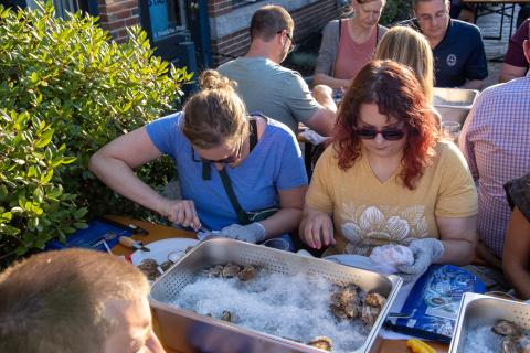 Two participants shuck oysters together at their table, with a bin of oysters on ice.
